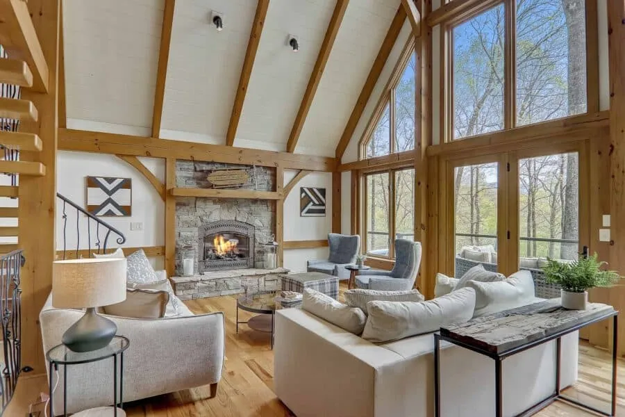 Cozy living room with a stone fireplace, vaulted wood-beamed ceiling, and large windows filling the space with natural light.