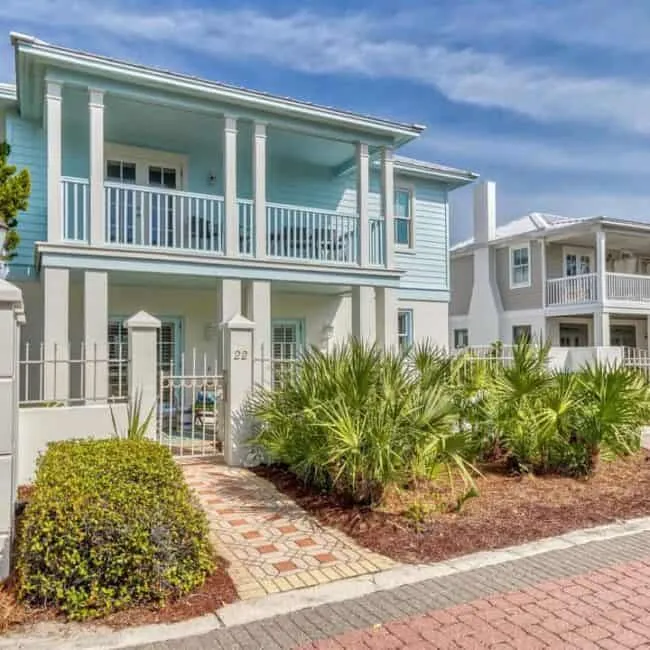 Two-story light-blue house with white railings and a second-floor balcony, front garden with palm plants.